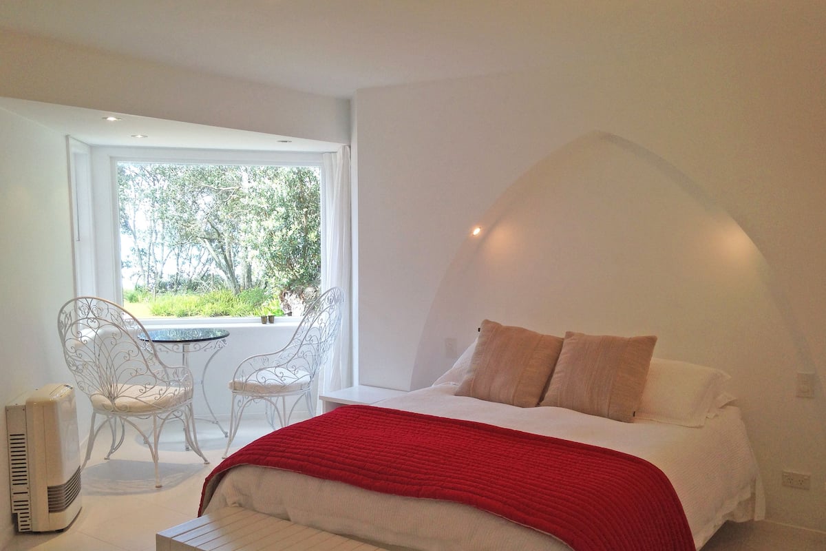 Een slaapkamer met een bed, een bankje, twee stoelen, een tafel en een raam met uitzicht op groen.