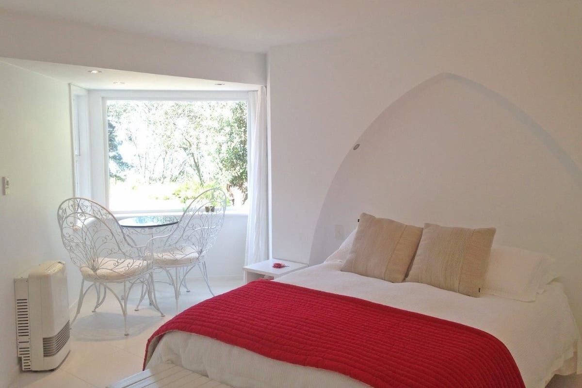 Een slaapkamer met een bed, een rode deken, een witte stoel en een klein tafeltje.