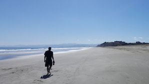 En person promenerar längs en bred, sandig strand under en klar himmel, med avlägsna berg i horisonten.