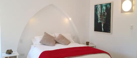 Une chambre avec un lit blanc, une couverture rouge, un banc blanc, une table de chevet ornée d’une petite figurine et un tableau accroché au mur.