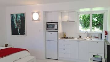 Une cuisine moderne et compacte, avec des meubles blancs, un four à micro-ondes et une cuisinière à gaz. On y trouve un lit avec une couverture rouge et une petite table de chevet. Une fenêtre laisse entrer la lumière naturelle et offre une vue sur la verdure environnante.