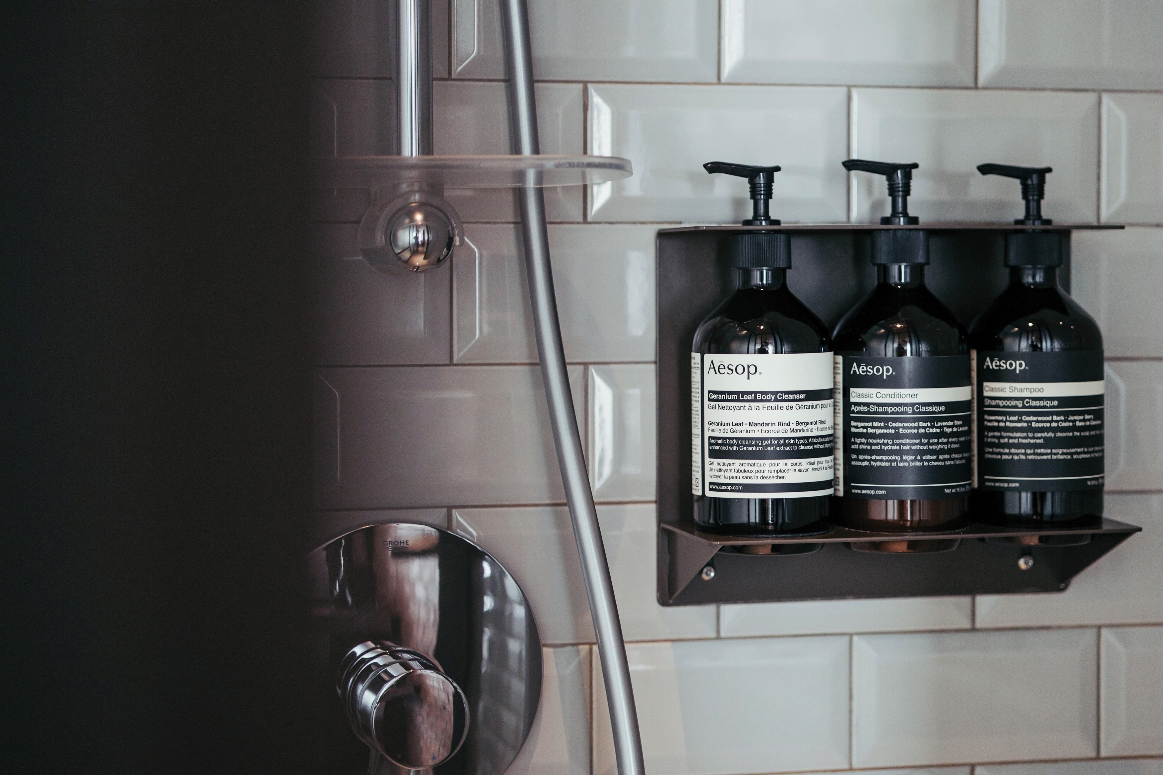 A bathroom with a shower, a sink, and three bottles of Aesop Classic Shampoo on a shelf.