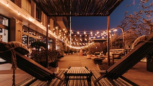 Un espacio de estar al aire libre con un banco de madera y sillones, luces colgantes y plantas en macetas.