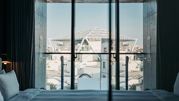 Habitación de hotel con un ventanal grande que ofrece vistas a un edificio moderno con una estructura de techo distintiva.