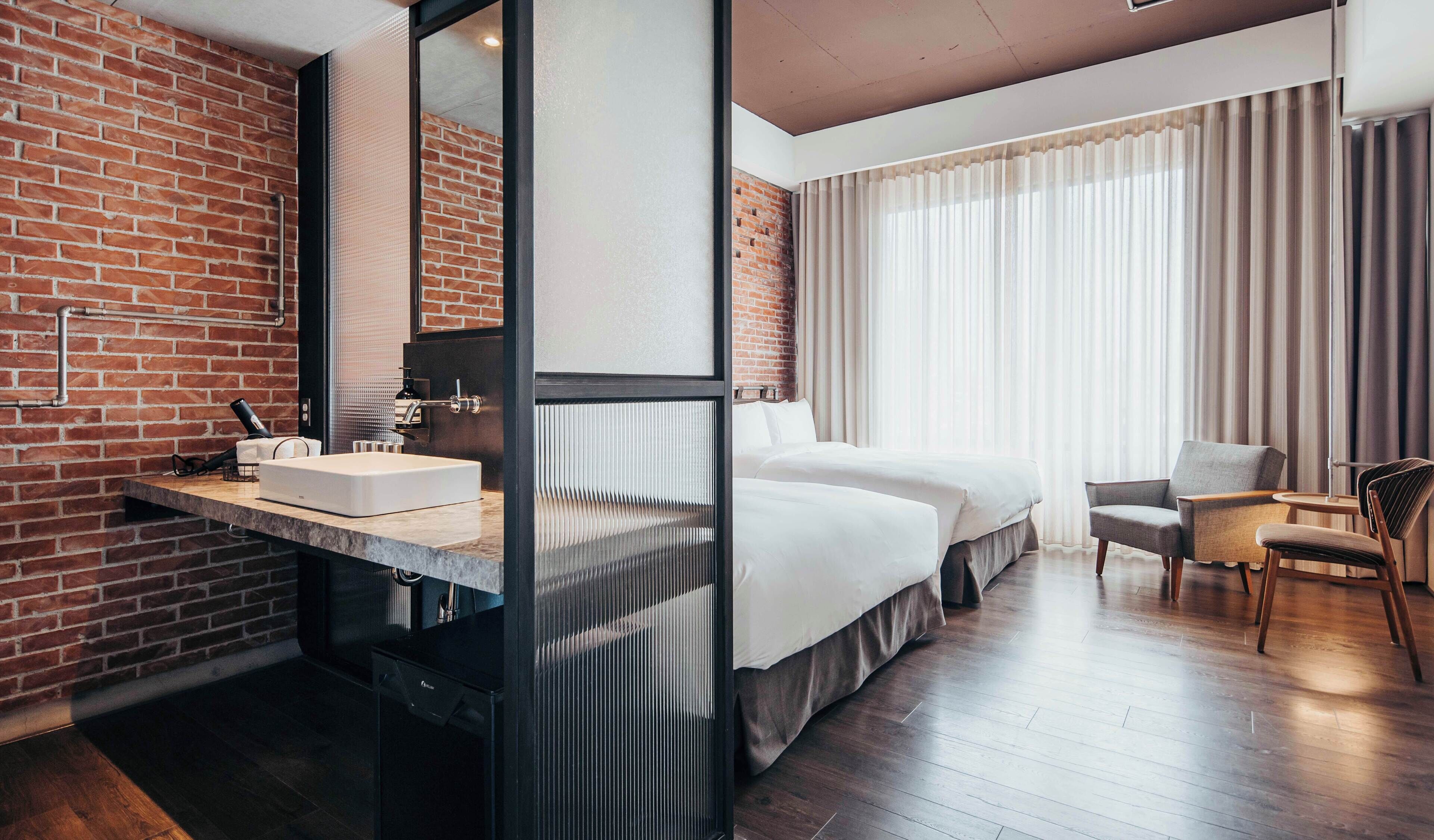 A modern hotel room with a brick wall, a large bed, a desk with a sink, and a chair.