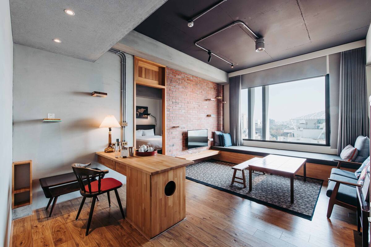 A modern hotel room with a wooden desk, a red chair, a sofa, a TV, and a large window offering a city view.