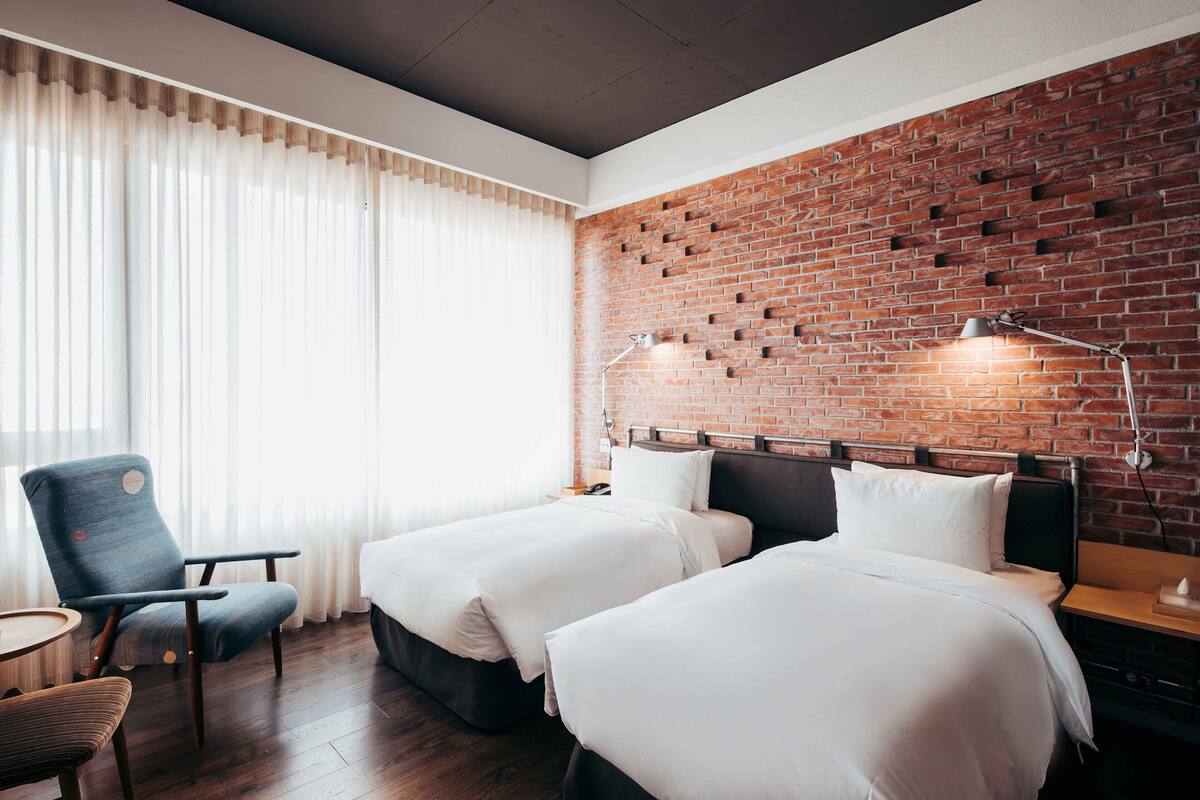 A hotel room with two beds, a chair, a table, and a brick wall.