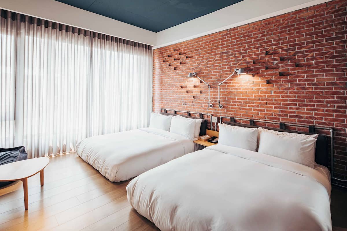 A hotel room with two beds, a desk, and a brick wall.