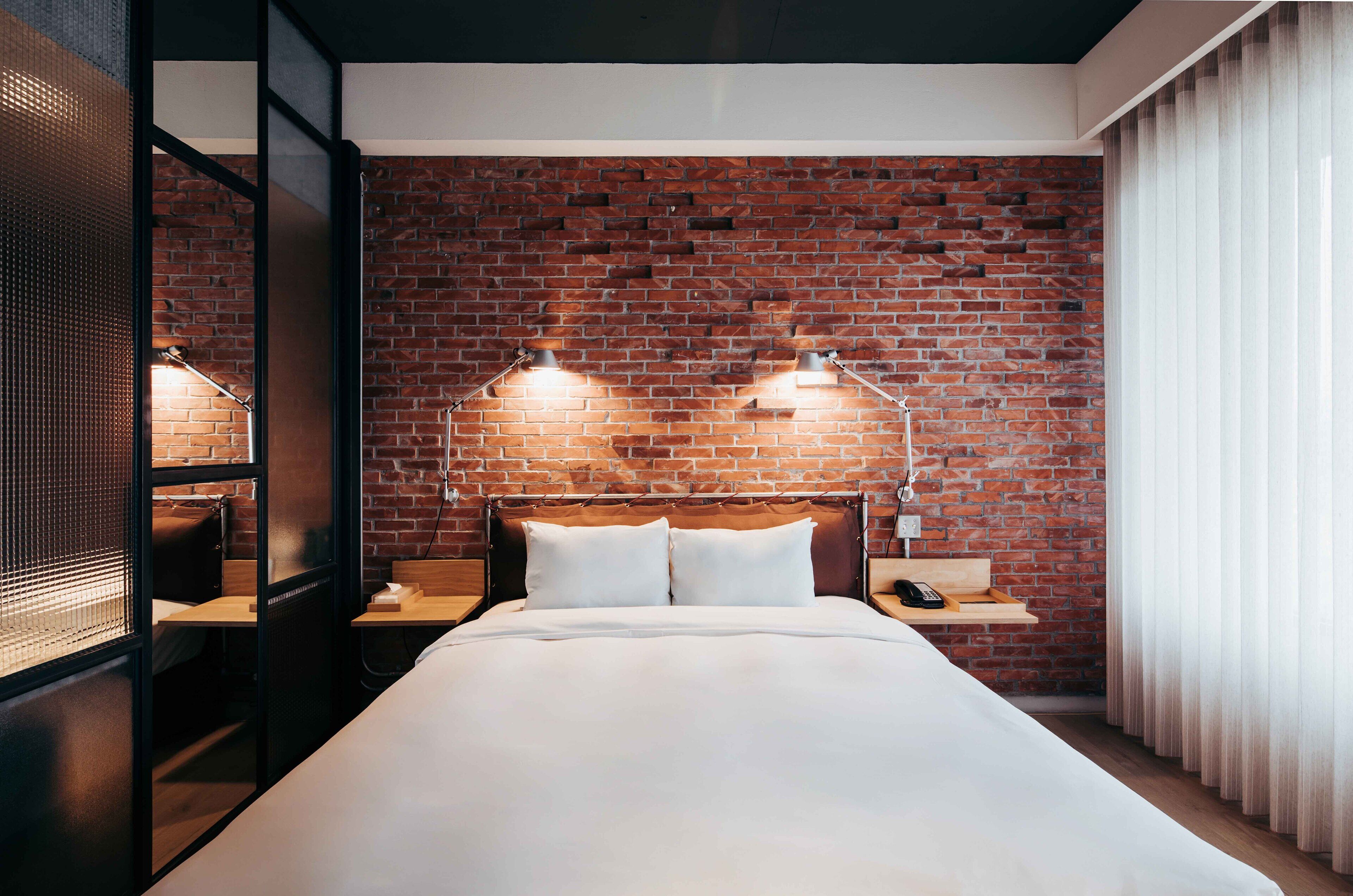 A hotel room with a brick wall, a bed with white linens, two bedside lamps, and a wooden nightstand with a phone.