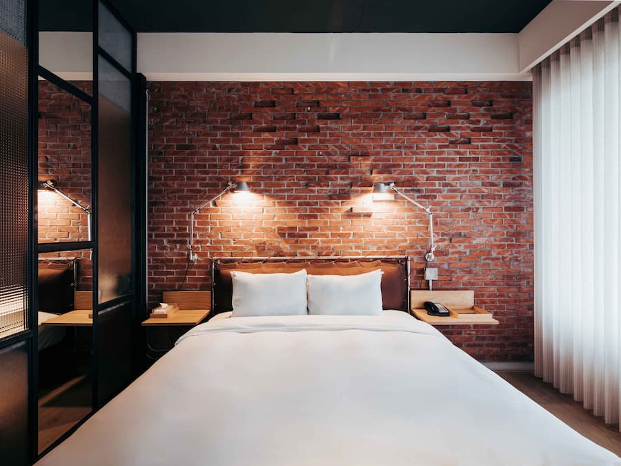 A hotel room with a brick wall, a bed with white linens, two bedside lamps, and a wooden nightstand with a phone.
