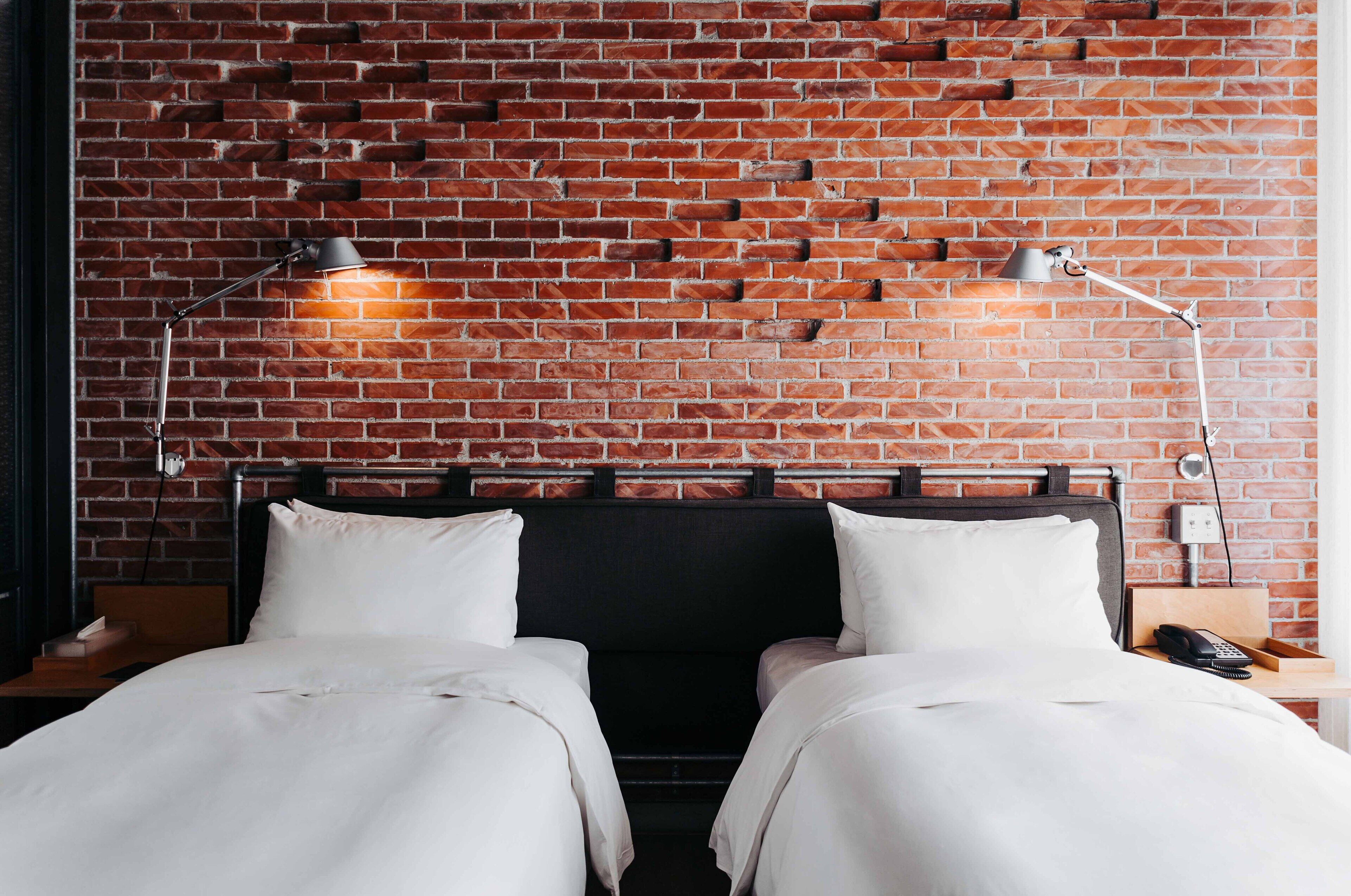 A hotel room with a brick wall, two beds with white bedding, a wooden headboard, and two bedside lamps.