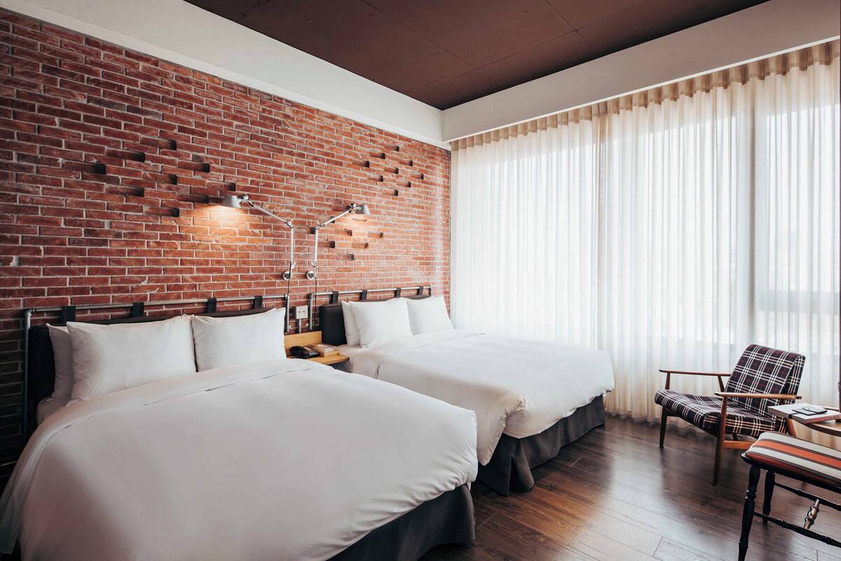 A hotel room with two beds, a brick wall, a large window with curtains, a chair, and a small table.