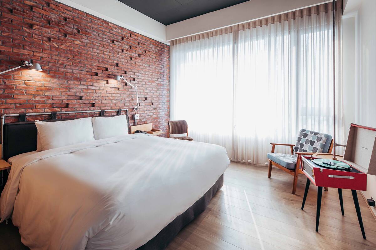 A hotel room with a large bed, a brick accent wall, a wooden floor, a desk with a red chair, and a window with sheer curtains.