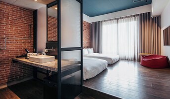 Une chambre d’hôtel avec un mur en briques, un grand miroir, un lavabo, un lit et un pouf rouge en forme de haricot.