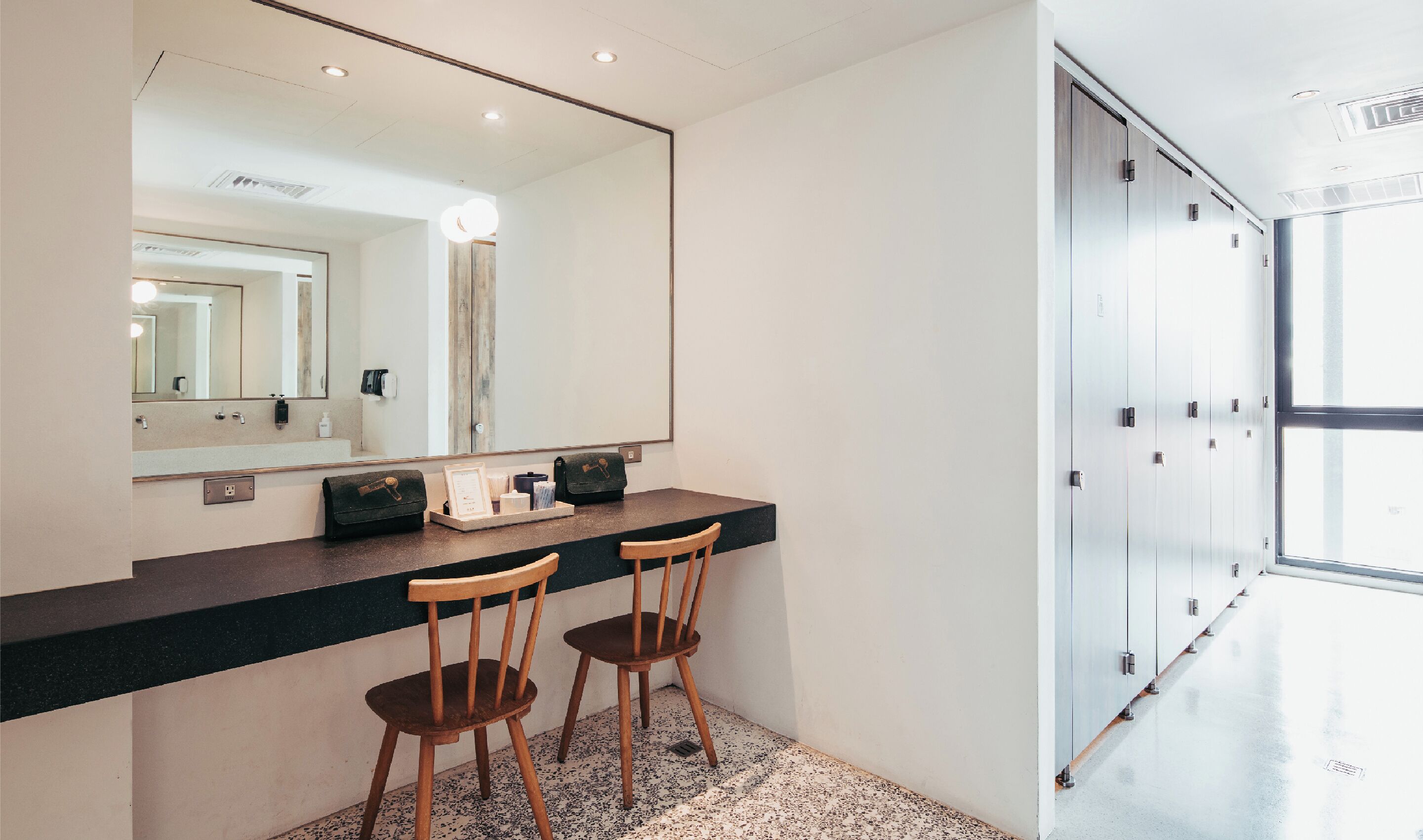 A modern bathroom with a large mirror, two wooden chairs, a vanity with a black countertop, and a glass door leading to a hallway.