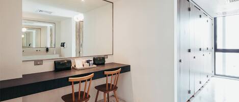 A modern bathroom with a large mirror, two wooden chairs, a vanity with a black countertop, and a glass door leading to a hallway.