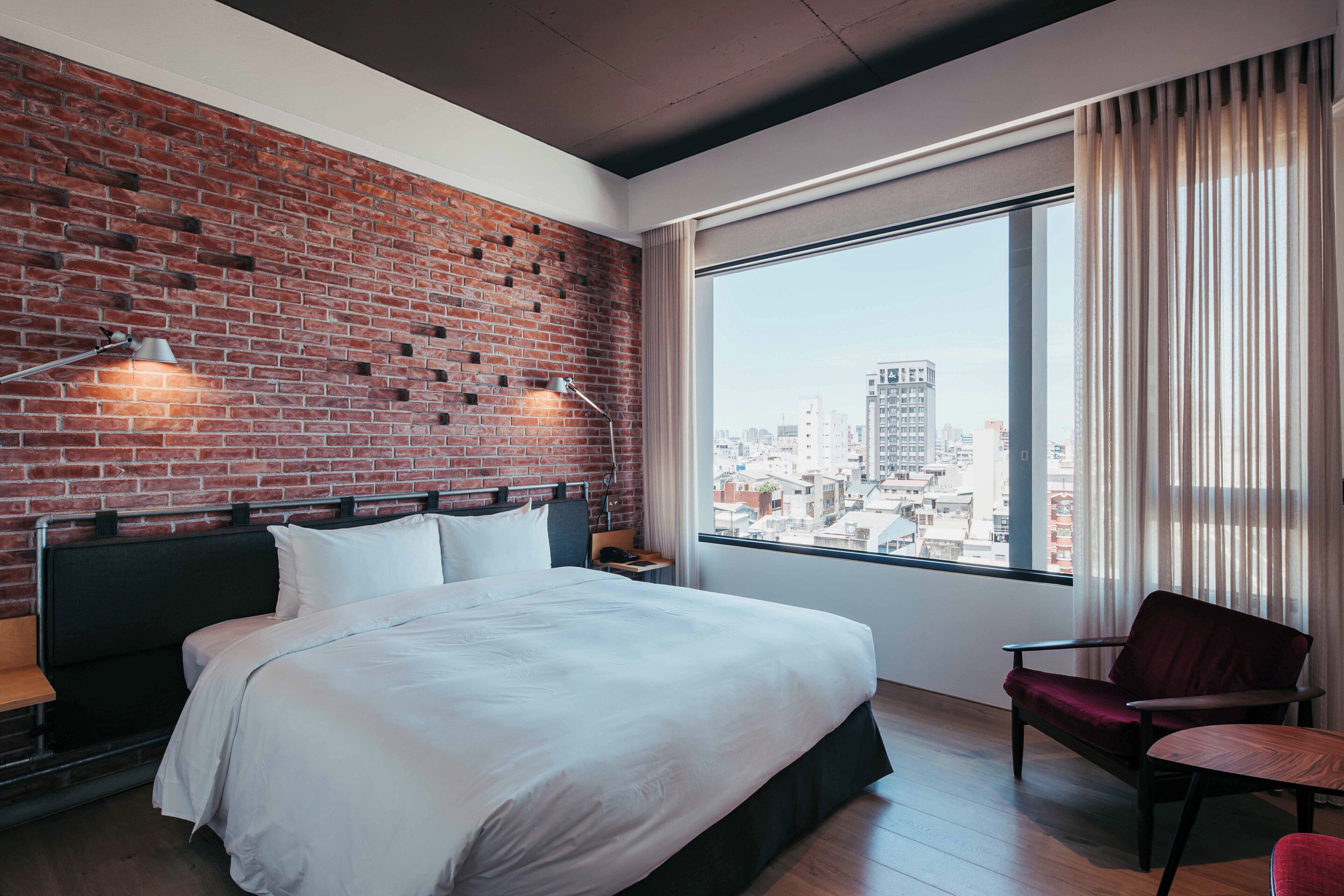 A hotel room with a brick wall, a large bed, a chair, and a view of the city.
