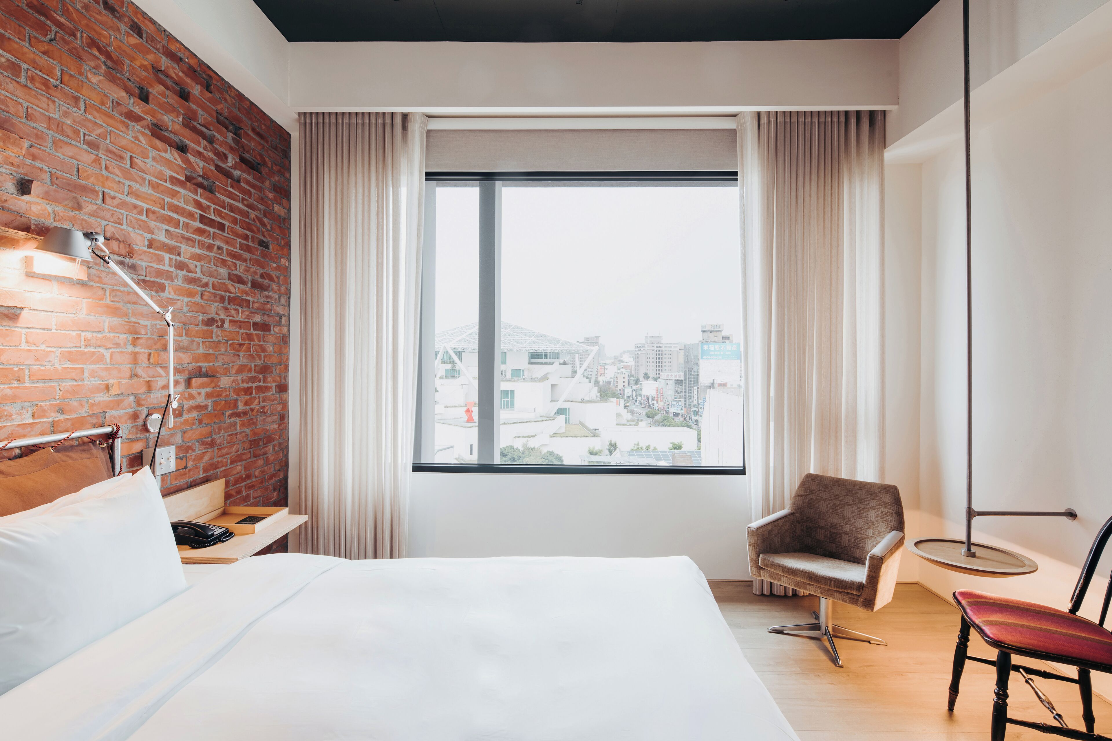 Een hotelkamer met een bakstenen muur, een groot raam met gordijnen, een bed, een stoel en een klein tafeltje.