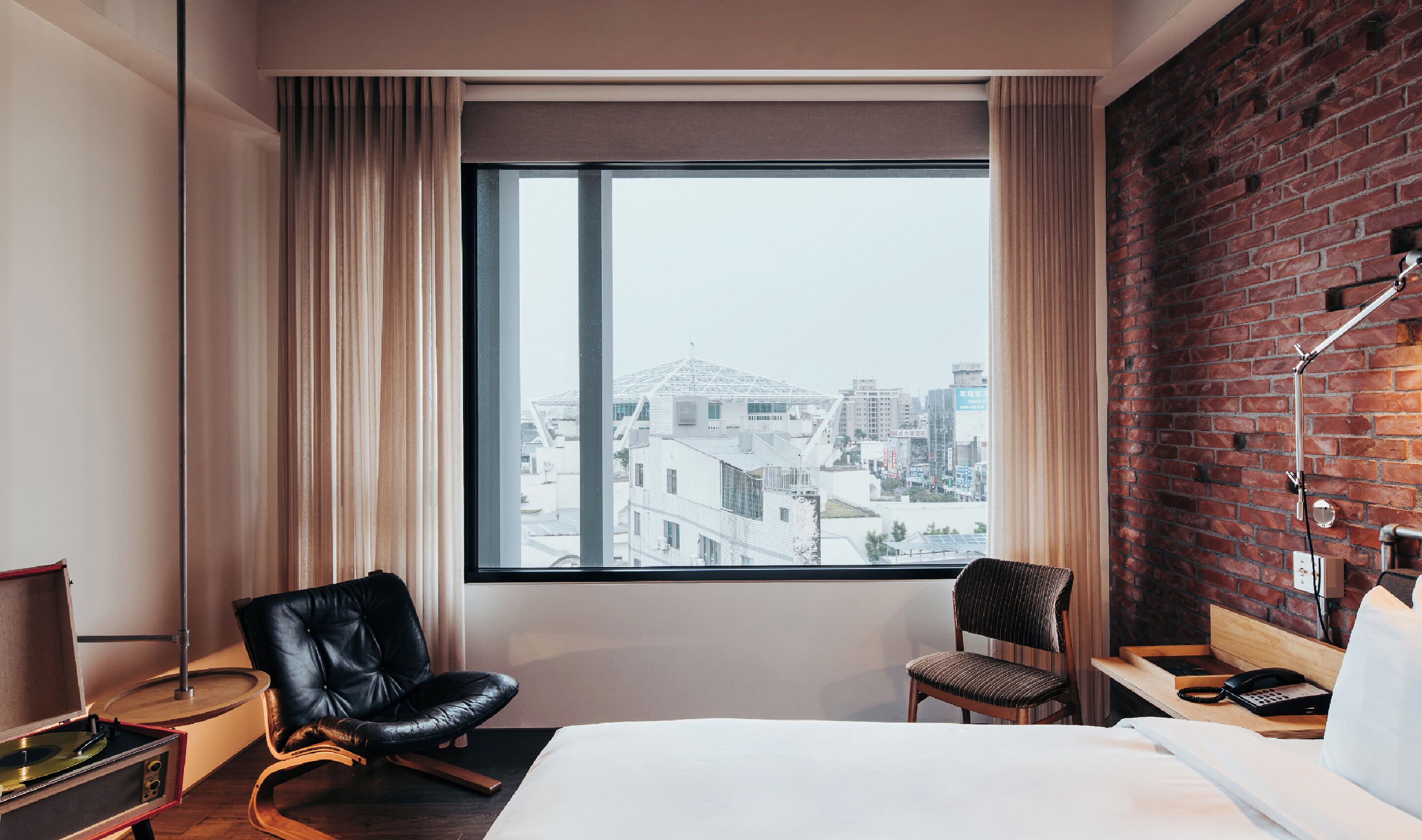 Habitación de hotel con una ventana grande, una cama, dos sillas, un escritorio con teléfono y una pared de ladrillo.
