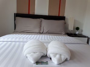 A neatly made bed with striped bedding, two folded towels, a bedside lamp, and a small potted plant.