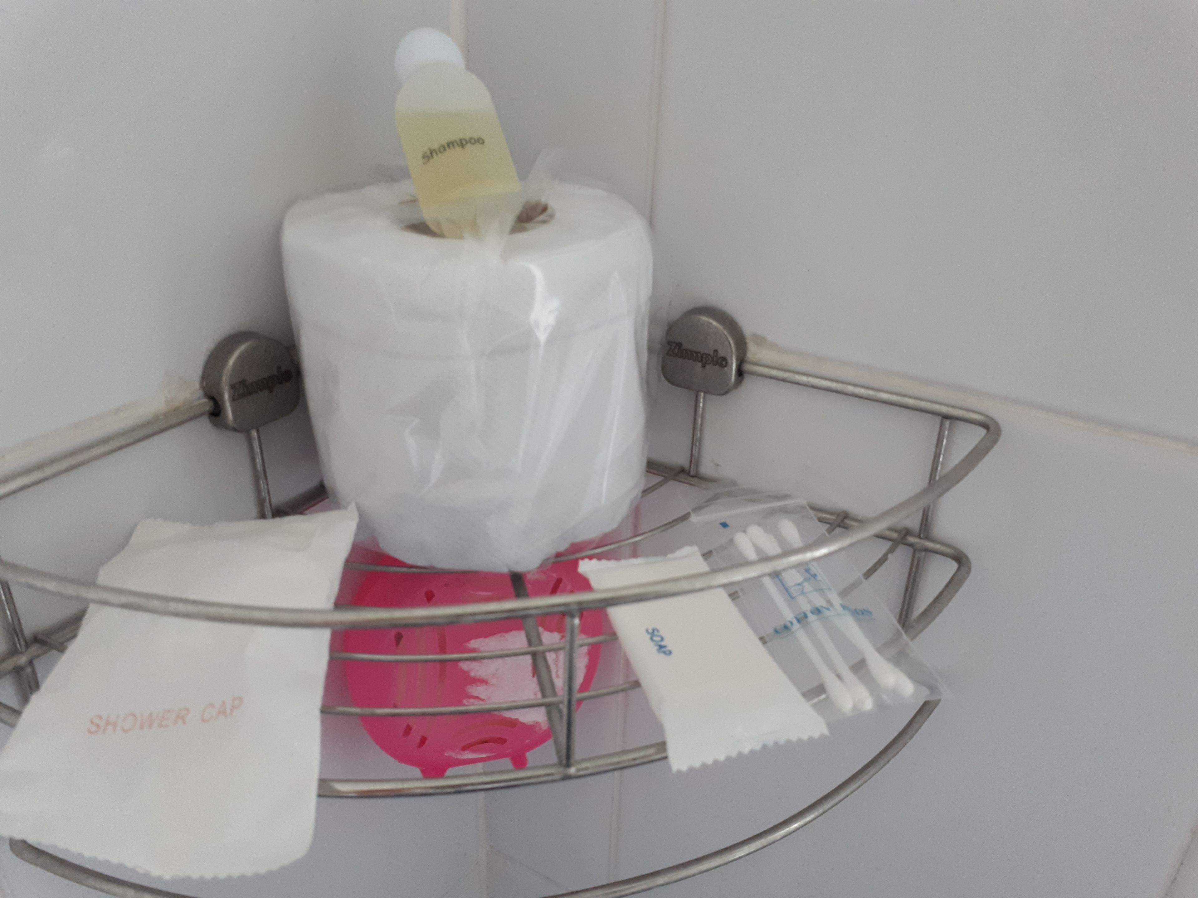 Ein Badezimmer mit einem Metall-Duschkorb, der eine Toilettenpapierrolle, eine Duschhaube und eine kleine Shampoo-Flasche enthält.