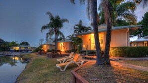 A resort with outdoor seating by a water body, palm trees, and a well-lit building at dusk. - Maroochy River Bungalows (Diddillibah)