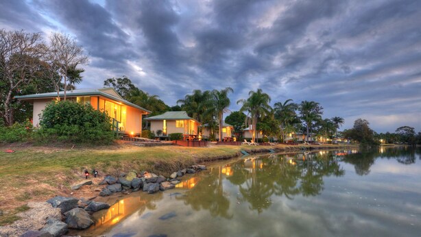 A lakeside property with multiple houses, a dock, and a calm water body reflecting the structures. - Maroochy River Bungalows (Diddillibah)