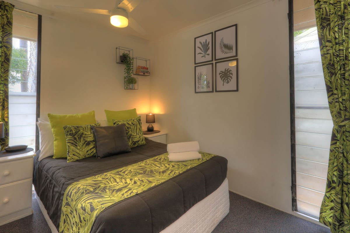 A bedroom with a bed, green patterned curtains, a bedside table with a lamp, and framed artwork on the wall.