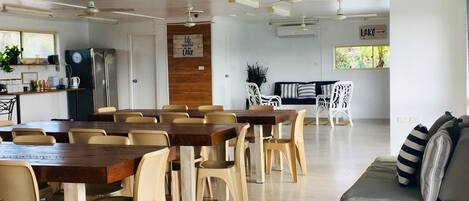 A spacious room with a dining area featuring wooden tables and beige chairs, a seating area with a sofa and cushions, and a kitchen with a refrigerator and a clock on the wall.