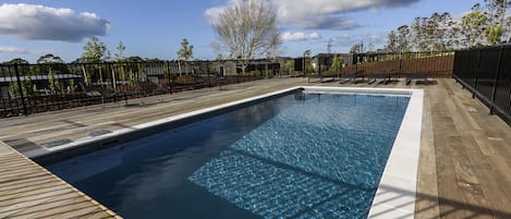 A rectangular swimming pool with a wooden deck surrounding it, a fence, and trees in the background.