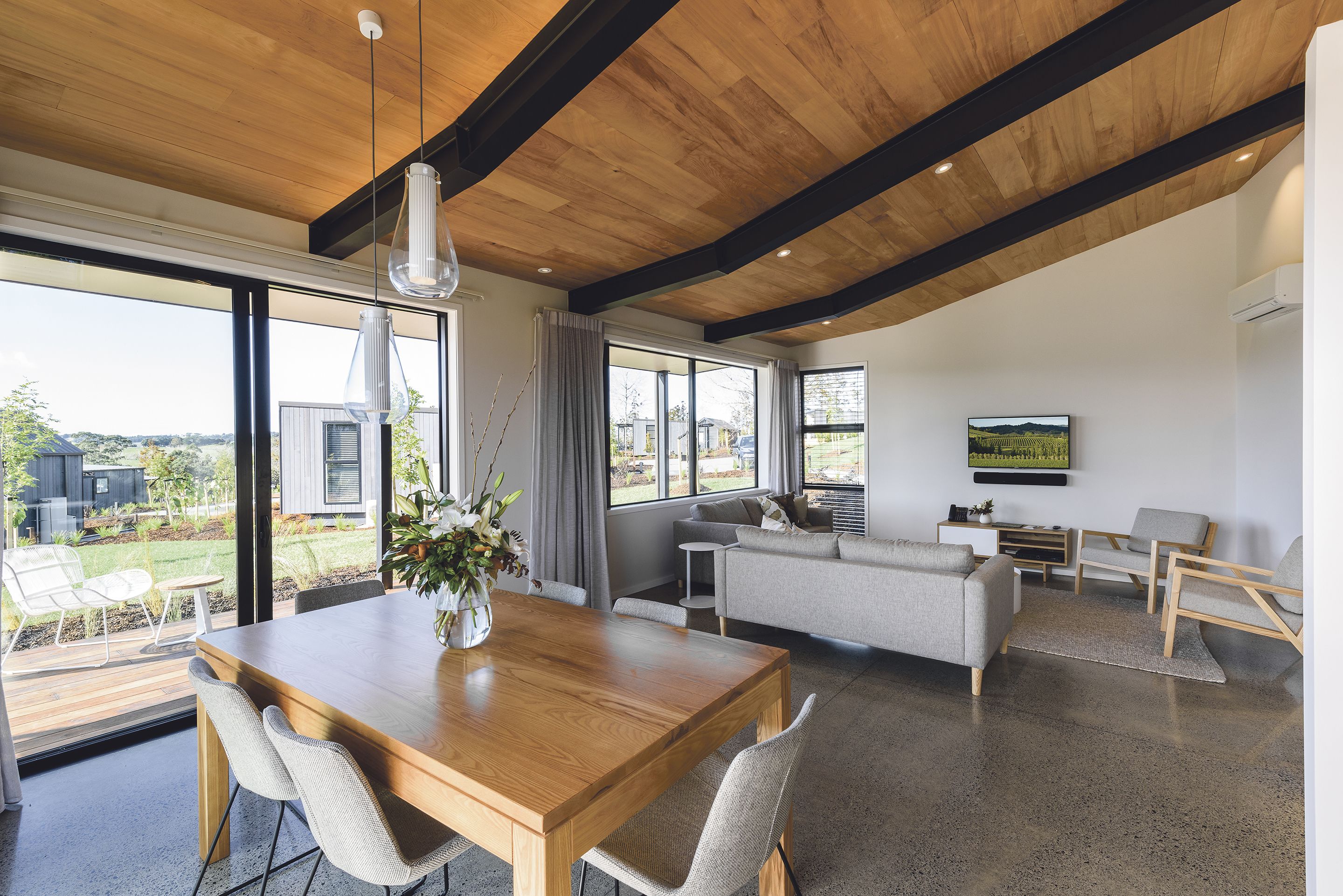 Een ruime woonkamer met een houten eettafel, grijze banken en een groot raam met uitzicht op de omgeving.