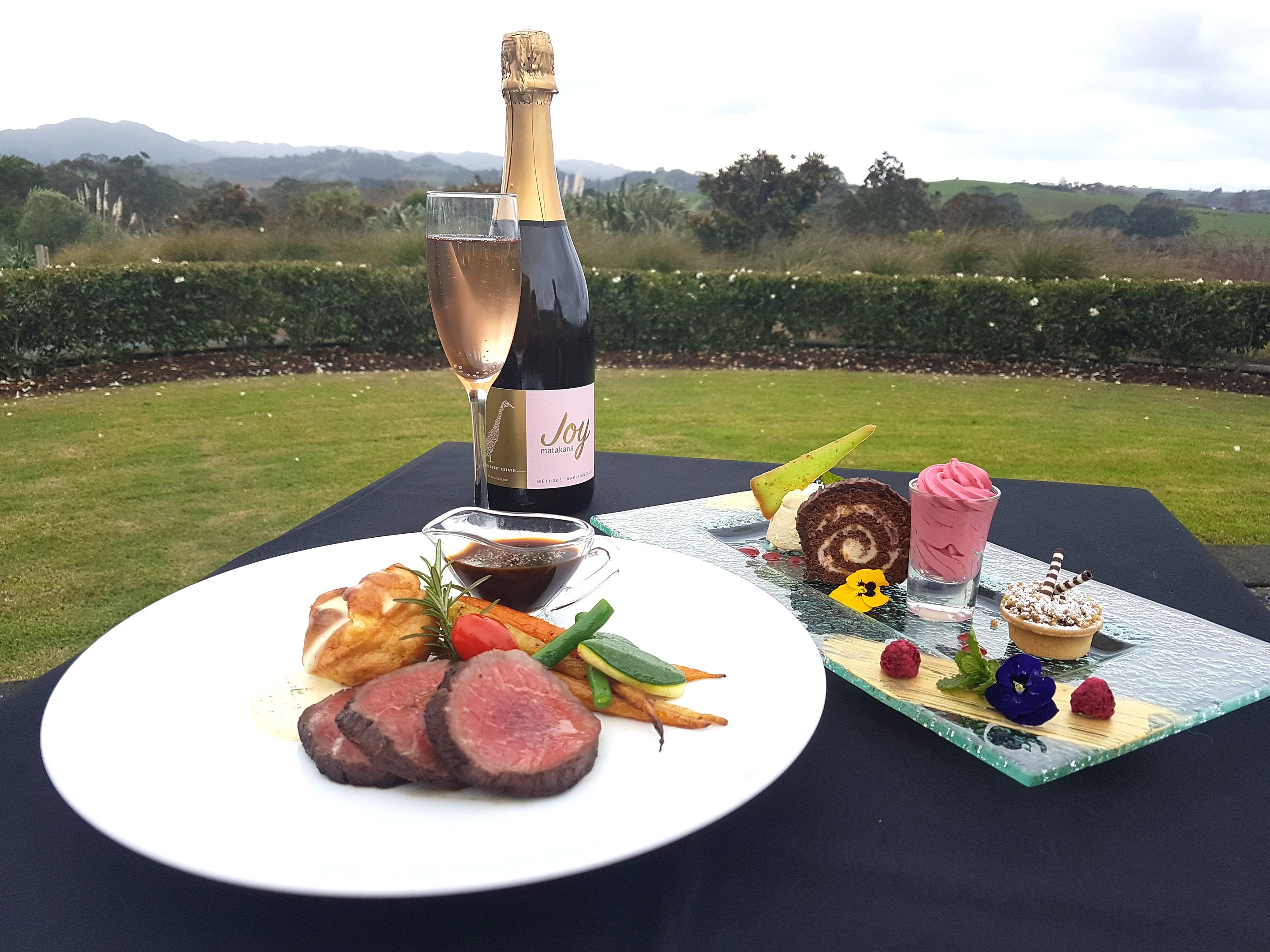 Un montaje de comida con carne, verduras y una botella de vino sobre una mesa con una vista panorámica al fondo.