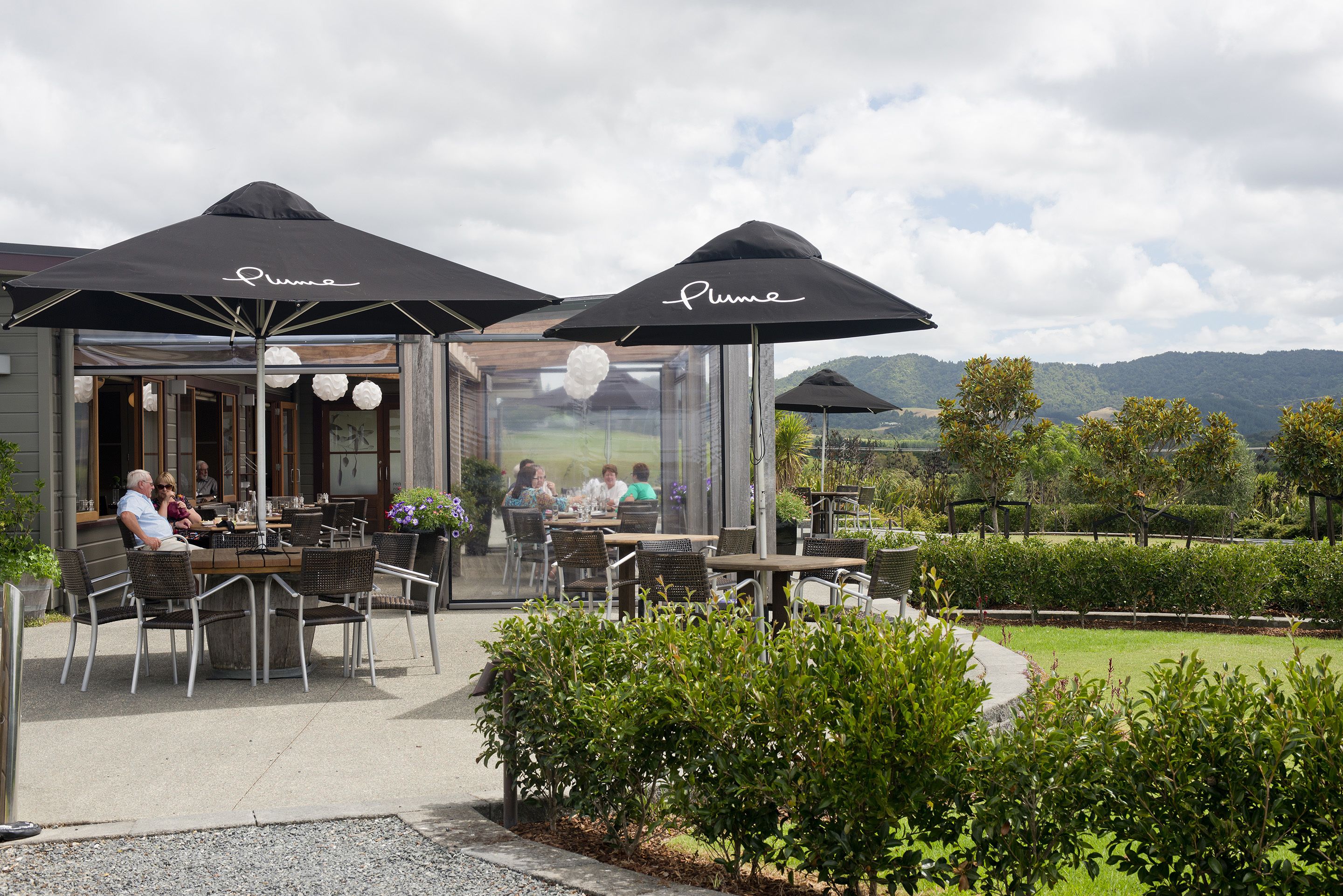 Un café en plein air avec des tables et des chaises sous des parasols, une paroi en verre et une vue sur le paysage.