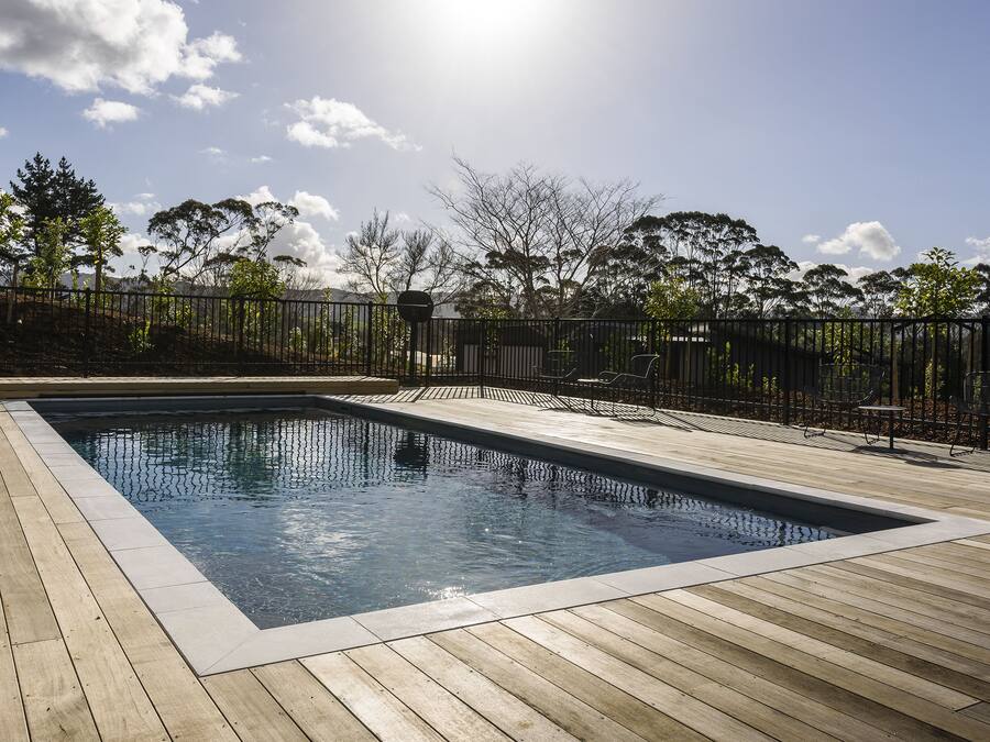 A rectangular swimming pool with a wooden deck, surrounded by a metal fence and trees.