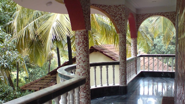 A curved staircase with white balustrades and tiled columns, leading to a balcony with a view of palm trees and a tiled roof. - Mama Mids Home (Morjim)