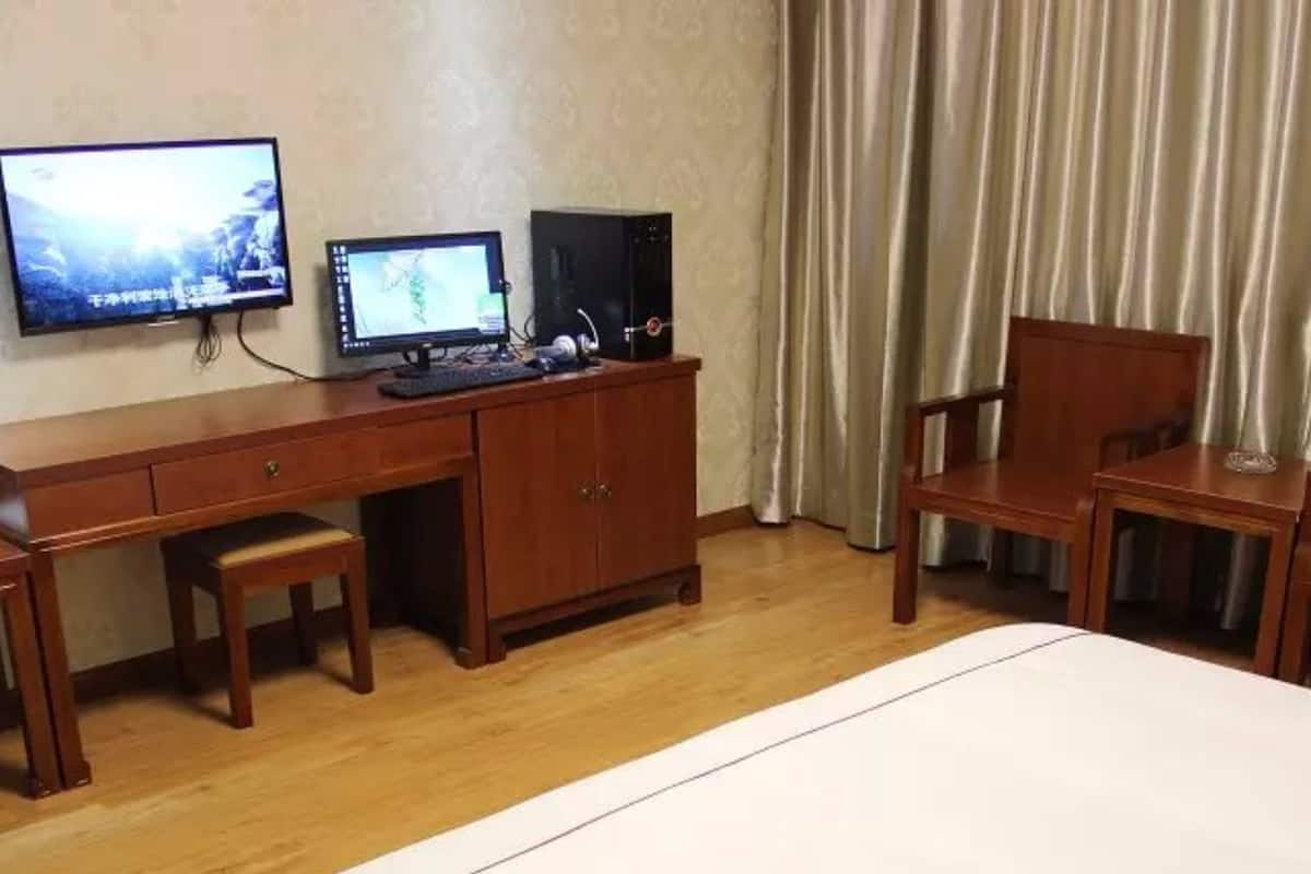 A hotel room with a bed, a desk with two monitors, a chair, and a curtain.