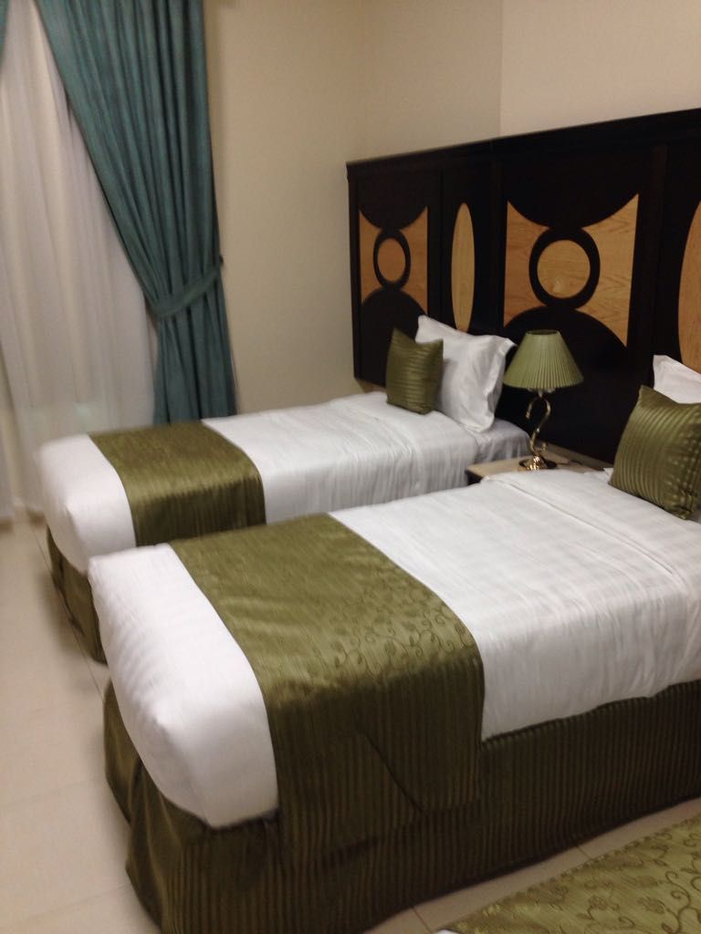 A hotel room with two beds, a headboard with a geometric pattern, a bedside table with a lamp, and a curtain.