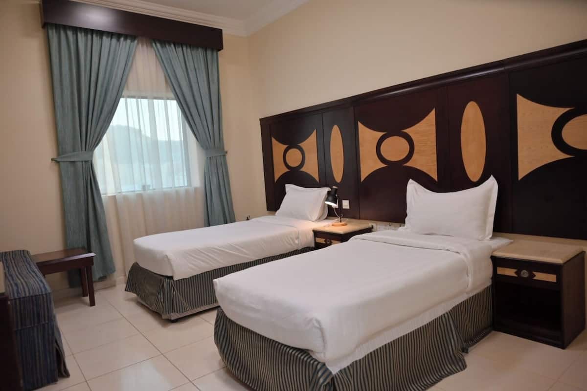 Habitación de hotel con dos camas individuales, una mesita de noche con lámpara, una pequeña mesa y un banco.