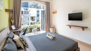 Habitación de hotel con cama, escritorio con computadora portátil, televisor montado en la pared y balcón con plantas y vistas a edificios.