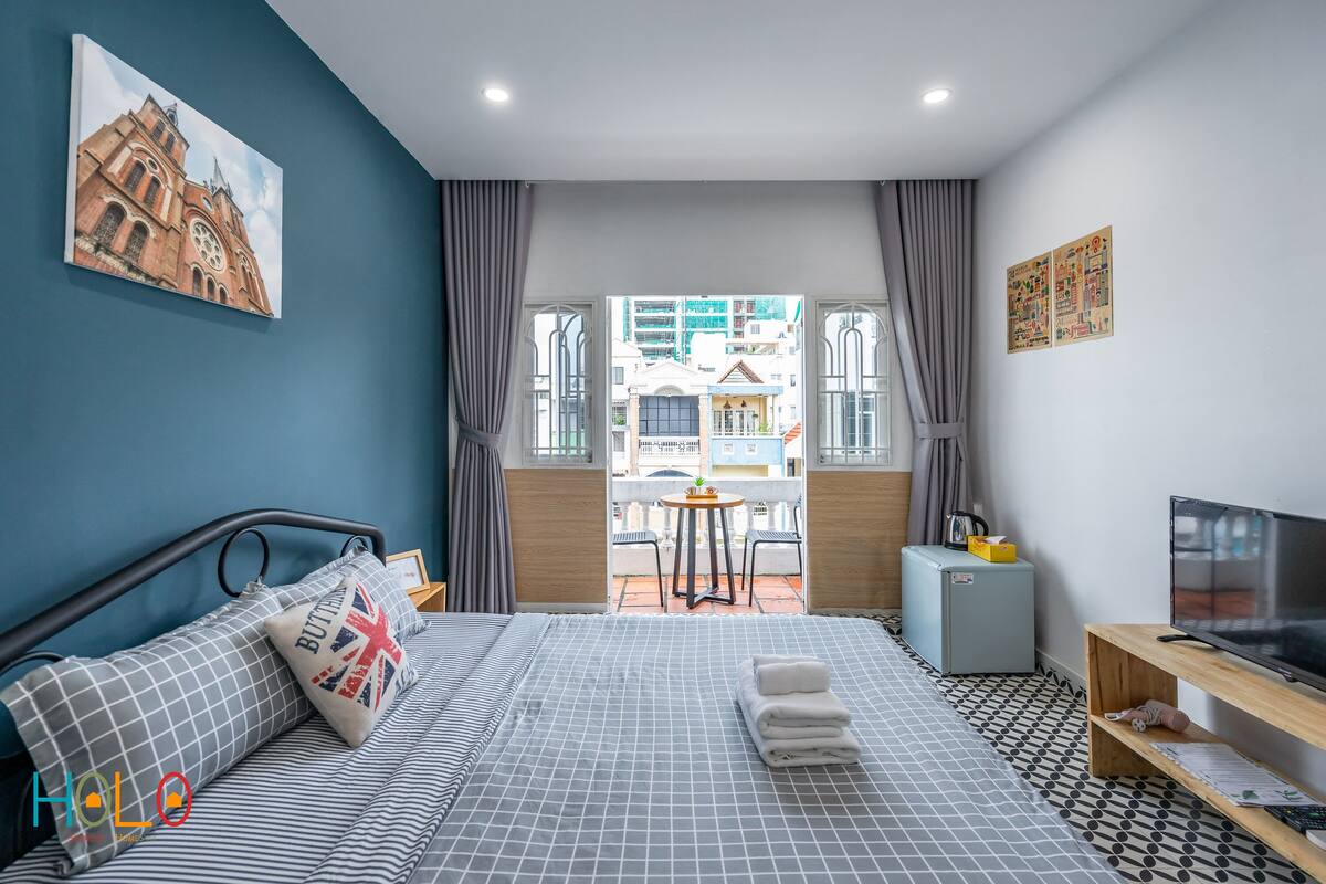 A modern hotel room with a bed, a TV, and a balcony with a view of buildings.