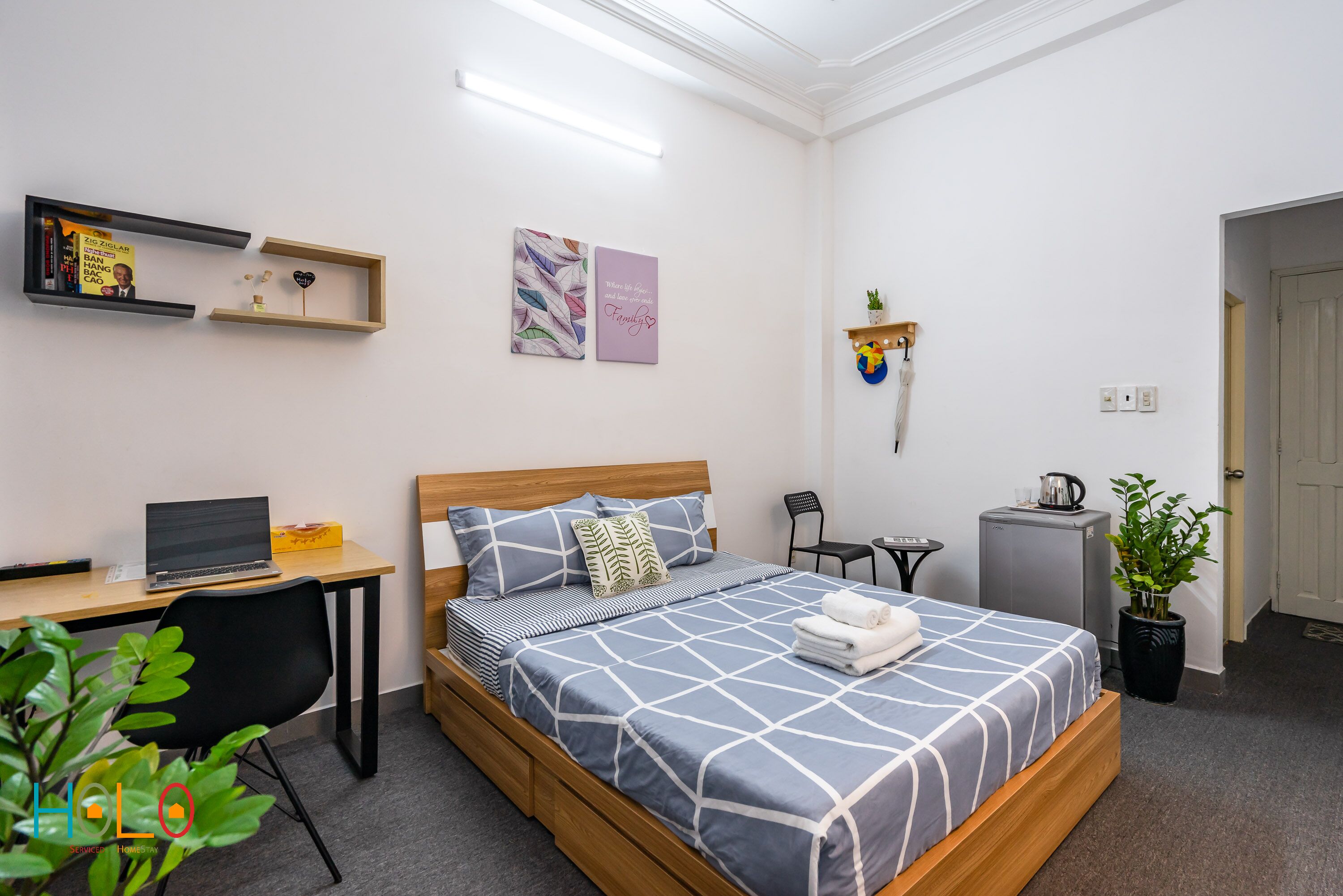 A neatly arranged bedroom with a bed, a desk, a chair, a small table, a potted plant, and a shelf with books and decorative items.