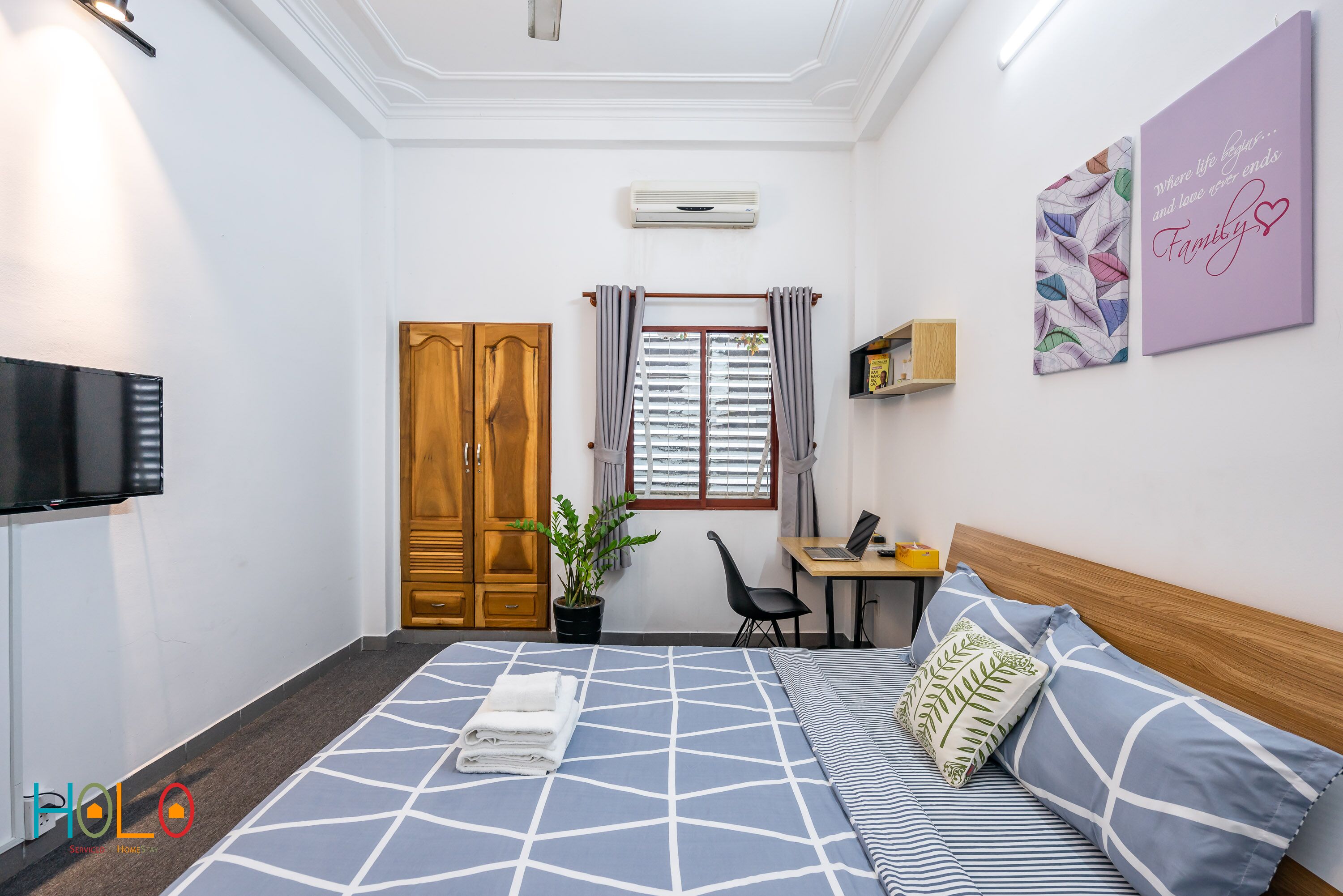 Quarto de hotel com cama, escrivaninha, cadeira, televisão, guarda-roupa e janela com cortinas.
