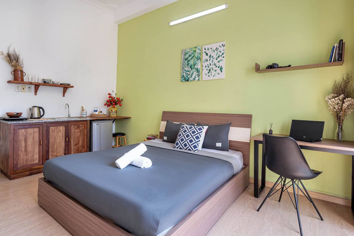 A bedroom with a bed, a desk, a chair, a kitchenette, and artwork on the walls.