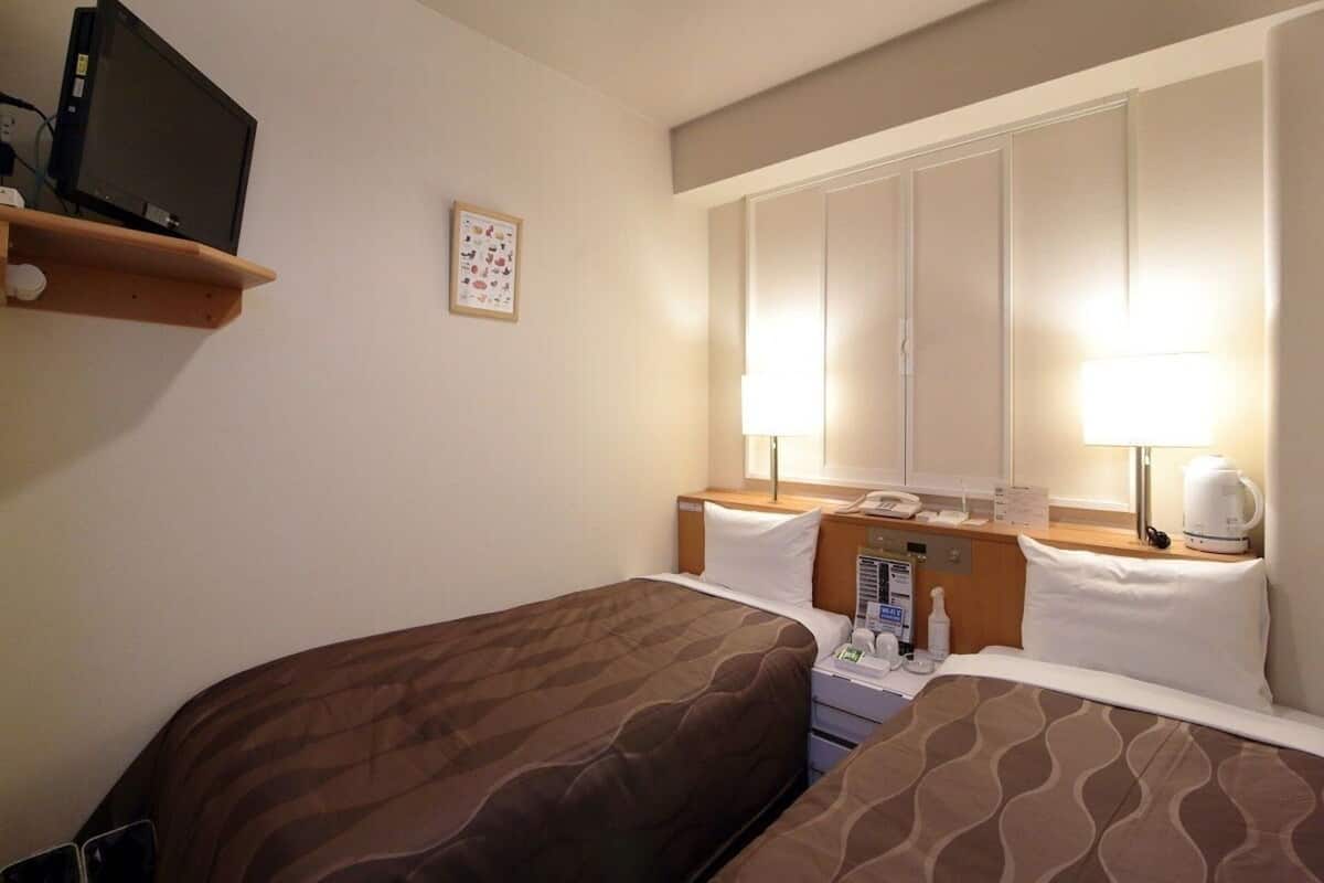 A hotel room with two beds, a TV on a shelf, a window with curtains, a bedside table with a lamp and a coffee maker, and a wall-mounted phone.