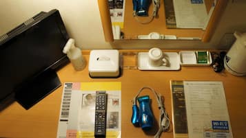 Un bureau dans une chambre d’hôtel avec un ordinateur portable, une bouteille de gel hydroalcoolique, une boîte de mouchoirs, une tasse, un petit plateau, une multiprise, un sèche-cheveux bleu, une télécommande et quelques documents imprimés.