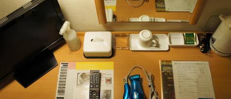 Escritorio de habitación de hotel con una computadora portátil, un dispensador de gel antibacterial, una caja de pañuelos, una taza, una pequeña bandeja, una regleta de enchufes, un secador de pelo azul, un control remoto y algunos documentos impresos.