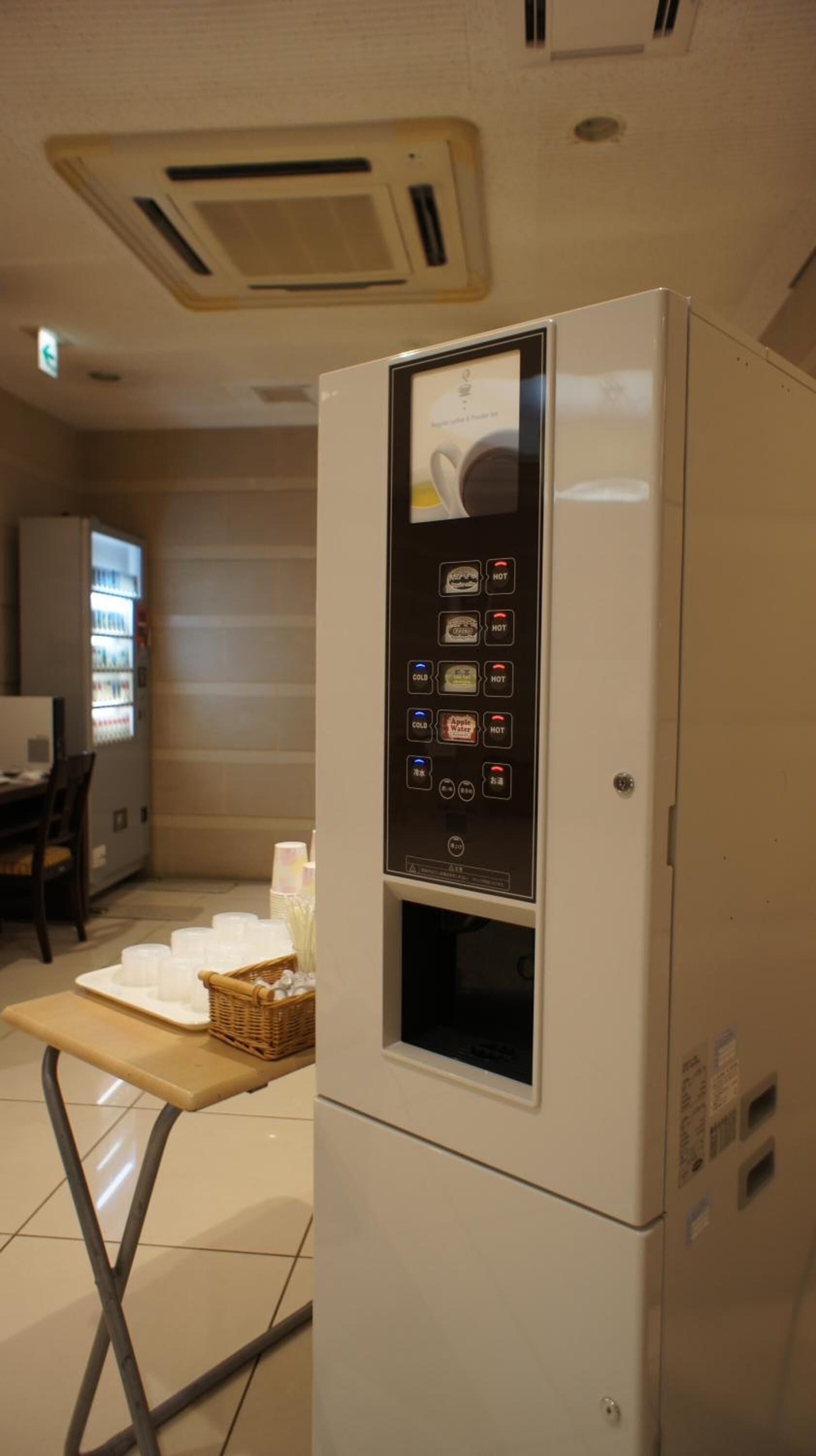 A self-service beverage dispenser with multiple drink options, a small table with cups and a basket, and a vending machine in a public area.