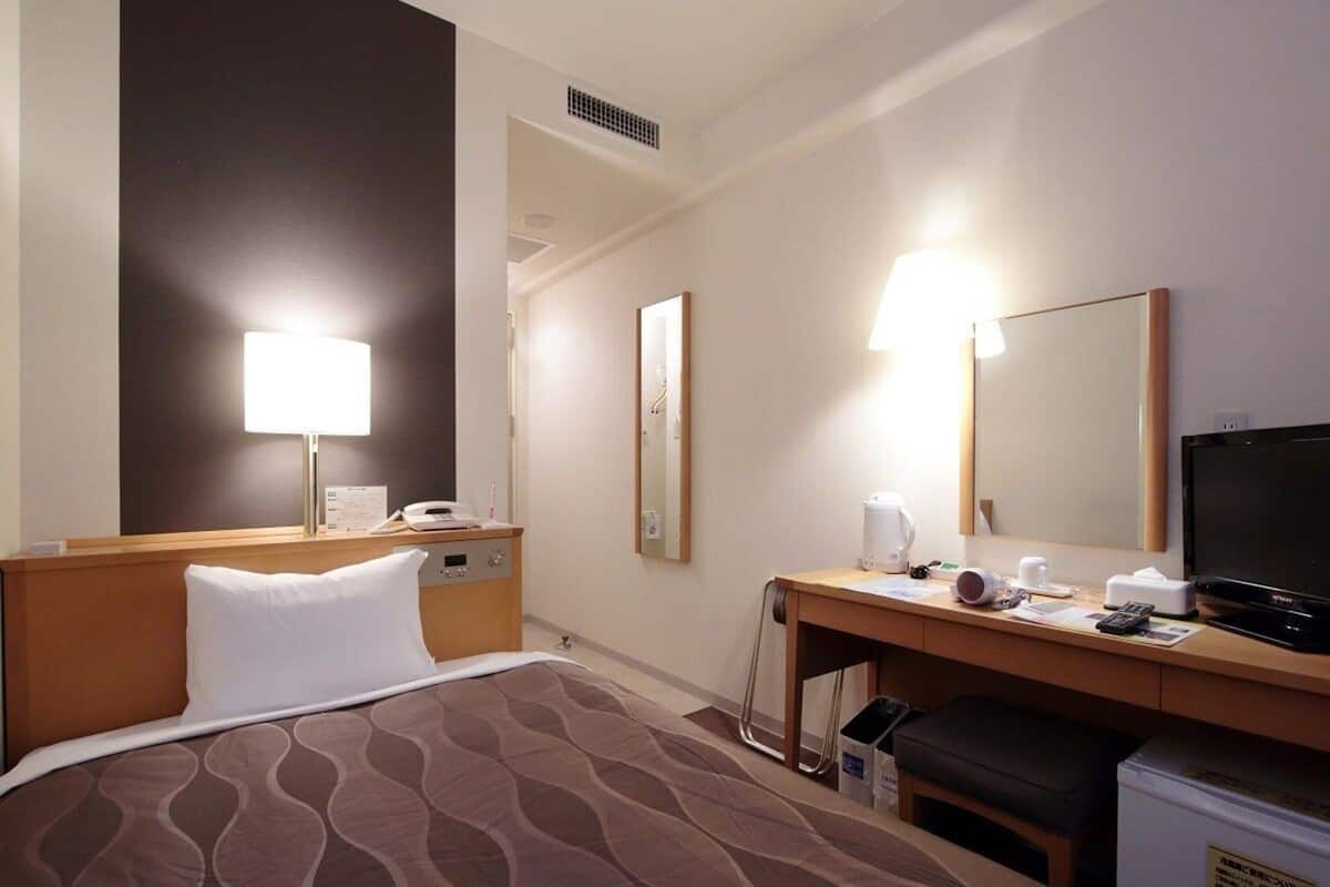 A hotel room with a bed, a desk with a computer, a television, and a refrigerator.