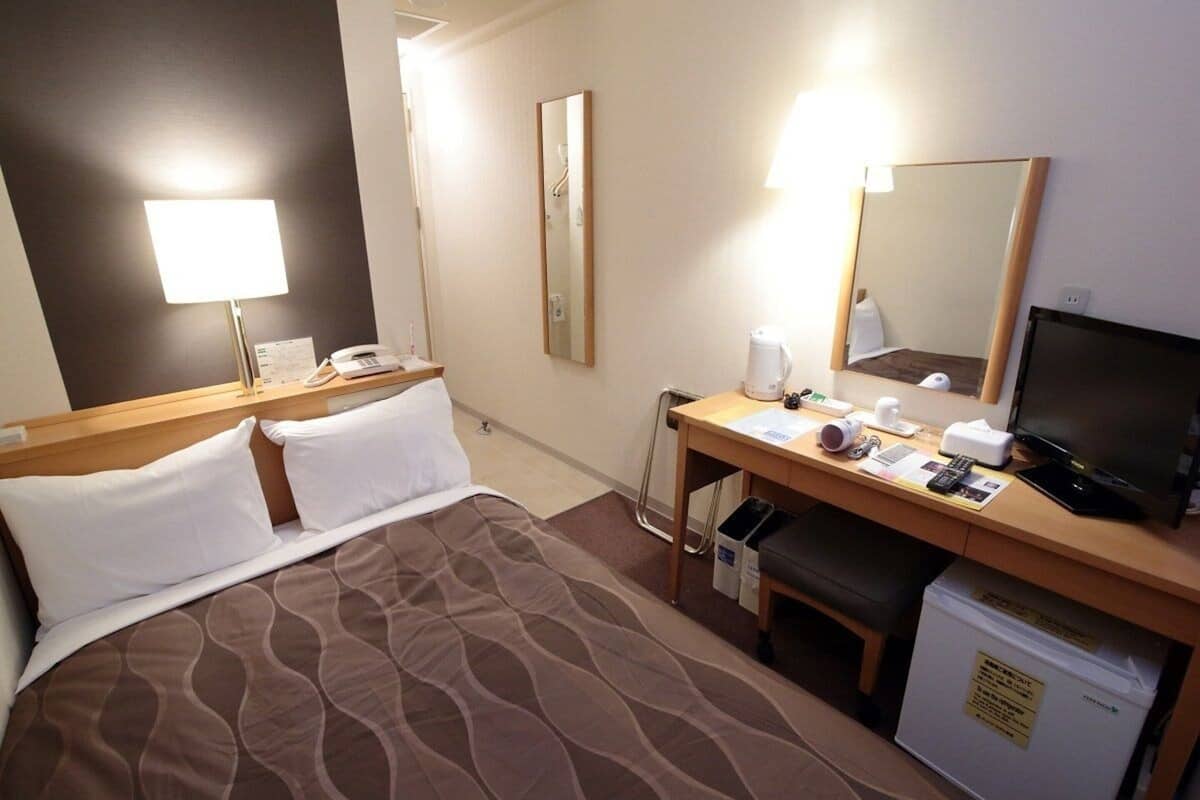 A hotel room with a bed, a desk with a computer, a lamp, a mirror, and a small refrigerator.