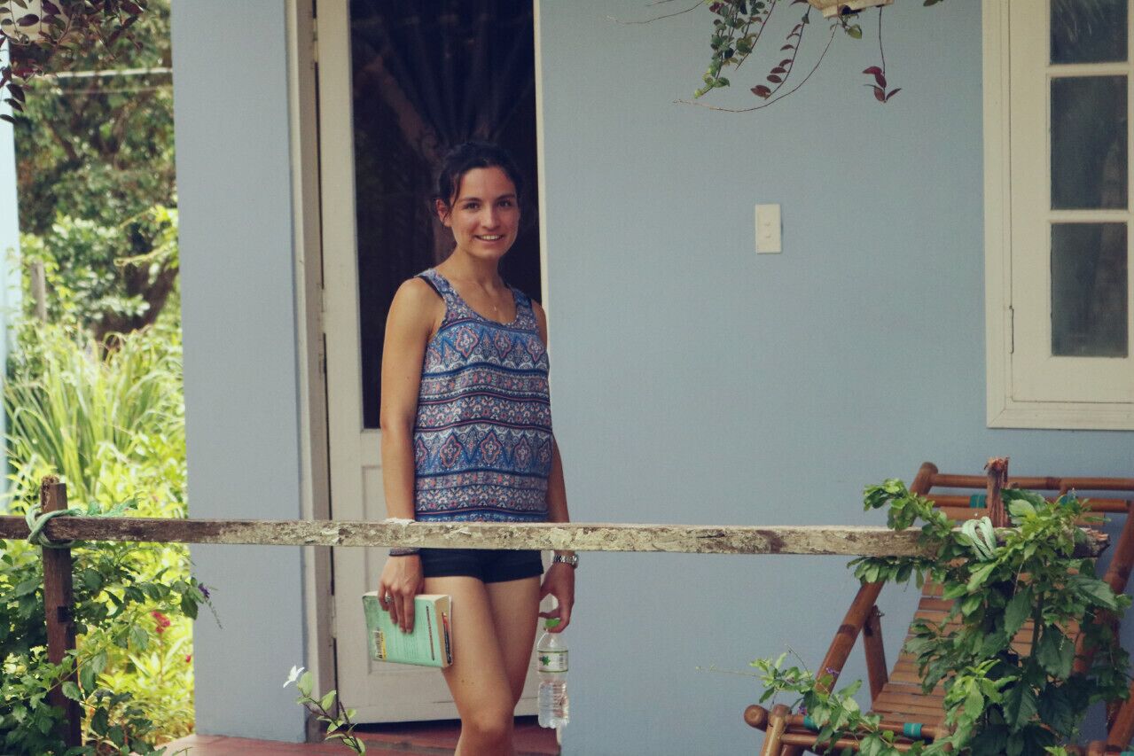 A person standing at the entrance of a building with a blue exterior, a wooden chair, and greenery around.